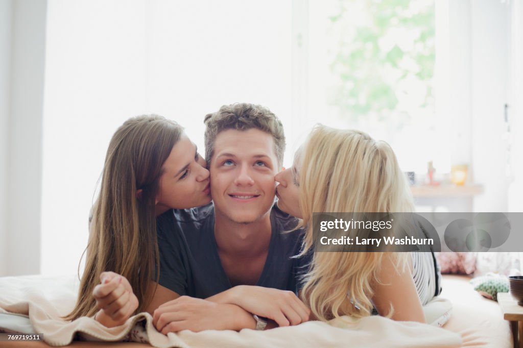 Young female friends kissing handsome man lying on bed at home