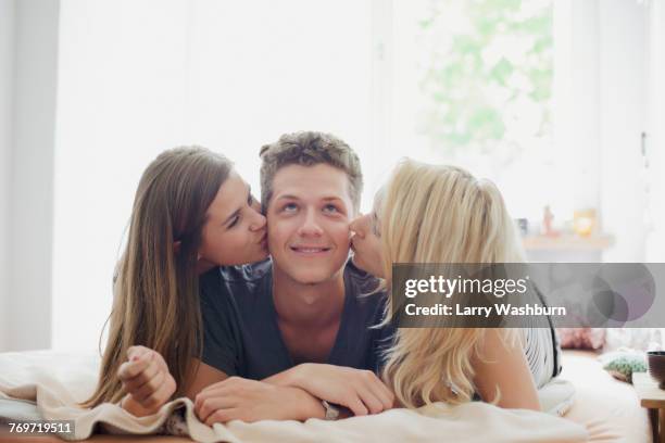 young female friends kissing handsome man lying on bed at home - three people in bed stock pictures, royalty-free photos & images