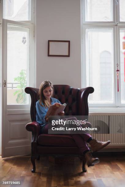 beautiful young female university student sitting on armchair while reading book at home - fashion woman floor cross legged stock pictures, royalty-free photos & images