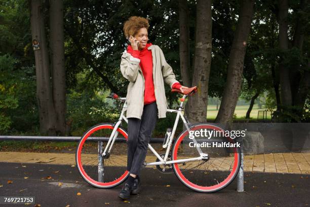 mid adult woman talking on mobile phone while standing with bicycle at park - berlin bike stock pictures, royalty-free photos & images