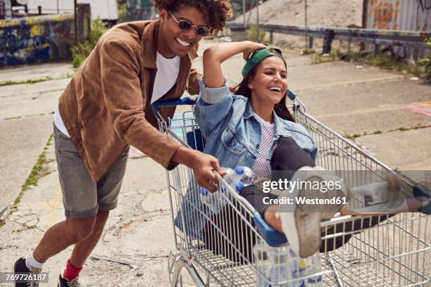 young man pushing girlfriend sitting in shopping cart - hipster summer fun stock-fotos und bilder