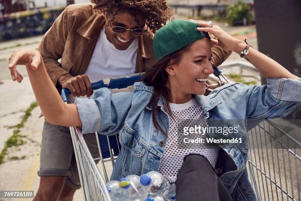 young man pushing girlfriend sitting in shopping cart - friend mischief stock-fotos und bilder