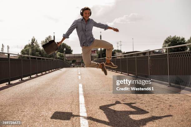 businessman with headphones and briefcase jumping for joy on a road - jumping for joy stockfoto's en -beelden