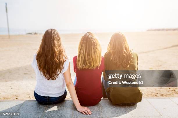 rear view of three female friends sitting at the beach - dreiviertel rückansicht stock-fotos und bilder