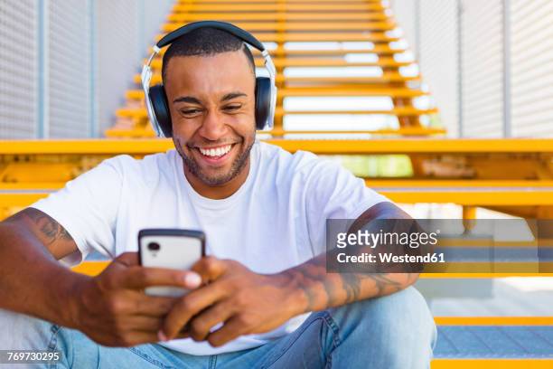 portrait of laughing young man with headphones sitting on stairs looking at smartphone - young man listening to music on smart phone outdoors stockfoto's en -beelden