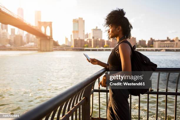 usa, new york city, brooklyn, woman with cell phone standing at the waterfront - african cityscape stockfoto's en -beelden