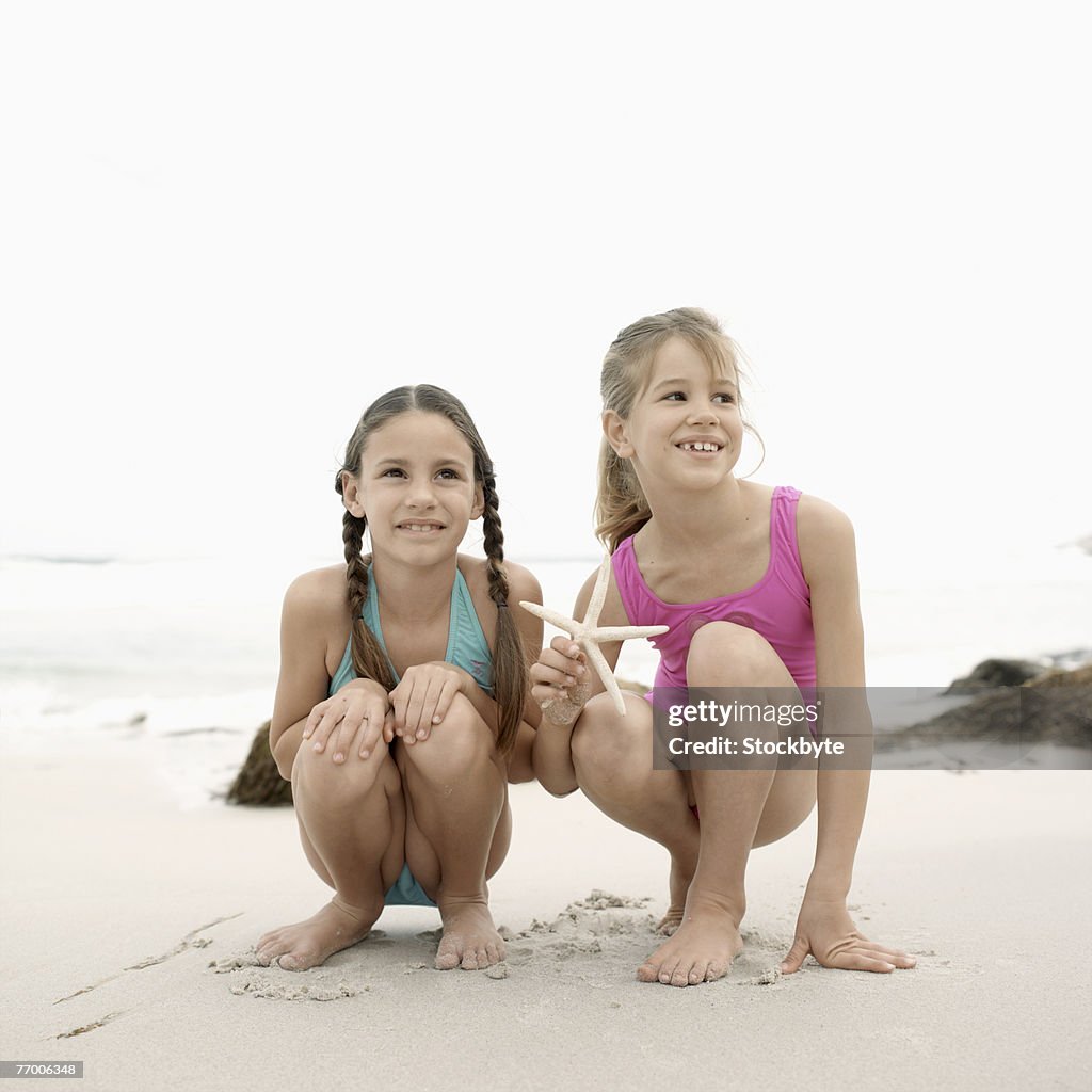 Two girls (9-10) in swimwear crouching on beach
