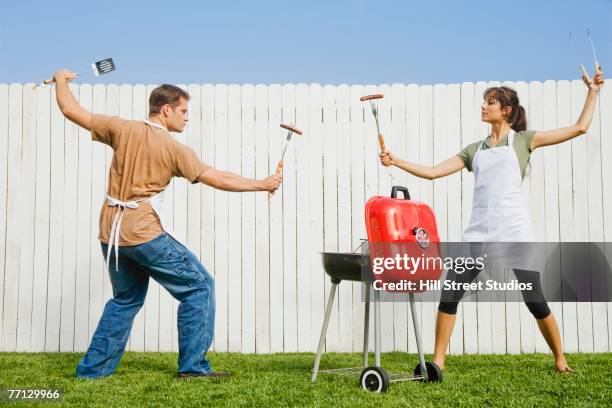 multi-ethnic couple in fighting stance with barbeque utensils - couple grilling stock-fotos und bilder