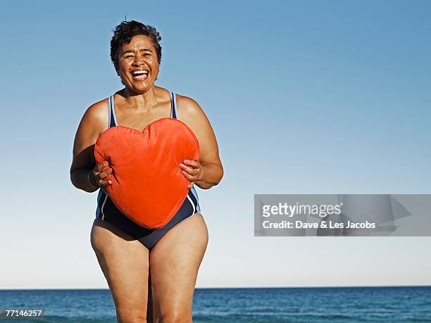 mixed race woman holding heart pillow at beach - old woman in swimsuit stock pictures, royalty-free photos & images