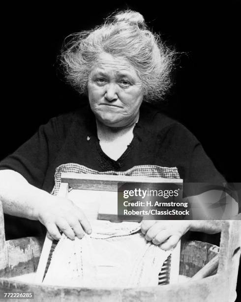 1930s DEPRESSION ERA SENIOR WOMAN SAD FACIAL EXPRESSION WASHING CLOTHES LAUNDRY ON SCRUBBING WASHBOARD LOOKING AT CAMERA