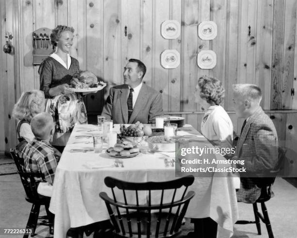 1950s MOTHER SERVING TURKEY ON PLATTER TO SMILING FAMILY AT TABLE IN KNOTTY PINE PANELED DINING ROOM