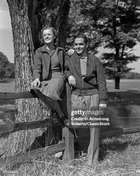 1940s TEEN COUPLE BOY GIRL HAPPY SMILING FULL LENGTH PORTRAIT BY WOODEN FENCE AND TREE