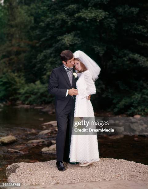 1960s FULL LENGTH PORTRAIT OF BRIDE AND GROOM STANDING ON ROCK BY CREEK OUTDOOR