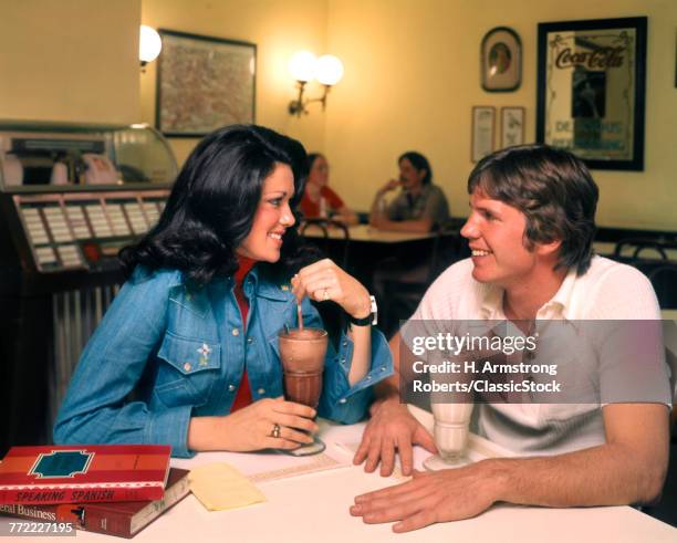 1970s YOUNG TEENAGE COUPLE SITTING IN SODA SHOP SHARING DRINKING MILKSHAKES TALKING JUKE BOX IN BACKGROUND