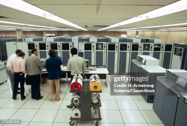 1970s SMALL GROUP OF MEN TECHNICIANS IN MAINFRAME COMPUTER ROOM WITH MULTIPLE MAGNETIC TAPE DRIVE MACHINES