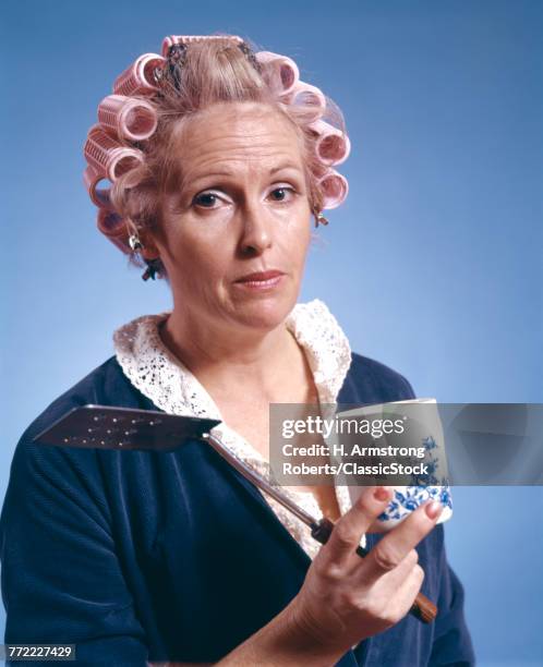 1970s MIDDLE AGED WOMAN WEARING CURLERS ROBE HOLDING EGG TURNER SPATULA COFFEE MUG HOUSEWIFE MORNING BREAKFAST