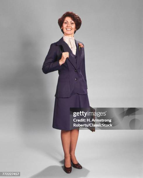 1970s CONFIDENT HAPPY SMILING BUSINESSWOMAN STANDING WEARING BLUE BUSINESS SUIT HOLDING BRIEFCASE LOOKING AT CAMERA