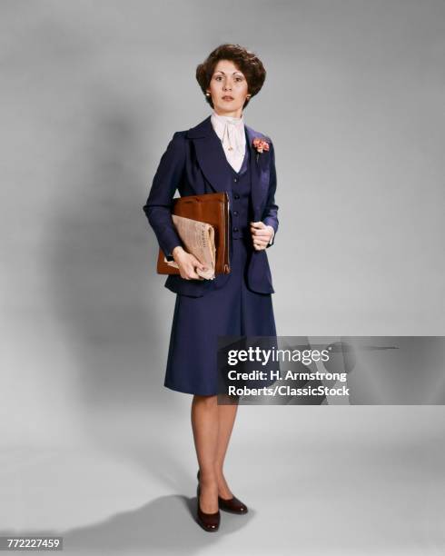 1970s BUSINESSWOMAN STANDING LOOKING AT CAMERA WITH SERIOUS FACIAL EXPRESSION HOLDING NEWSPAPER AND BRIEFCASE WEARING BLUE SUIT