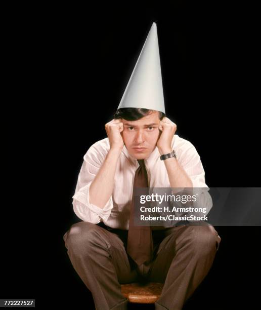 1960s UNHAPPY YOUNG MAN SITTING ON STOOL WEARING DUNCE CAP LOOKING AT CAMERA