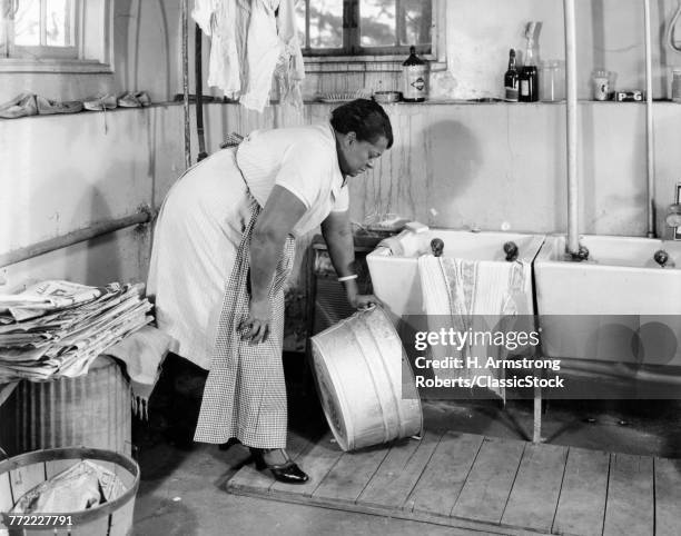 1940s AFRICAN AMERICAN OVERWEIGHT WOMAN STOOPING IN LAUNDRY ROOM PICKING UP GALVANIZED WASH TUB