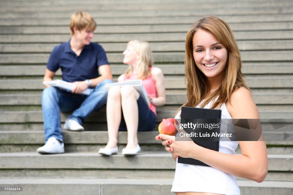 Young student on campus with book and apple. Other students in background