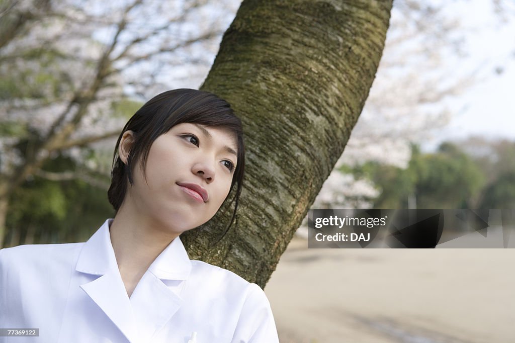 Woman in lab coat leaning on a tree trunk, front view, Japan