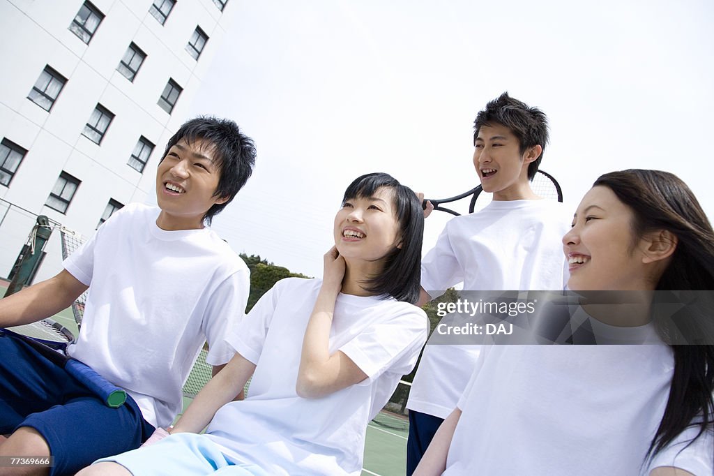 Four teenagers sitting on bench in tennis court, holding rackets