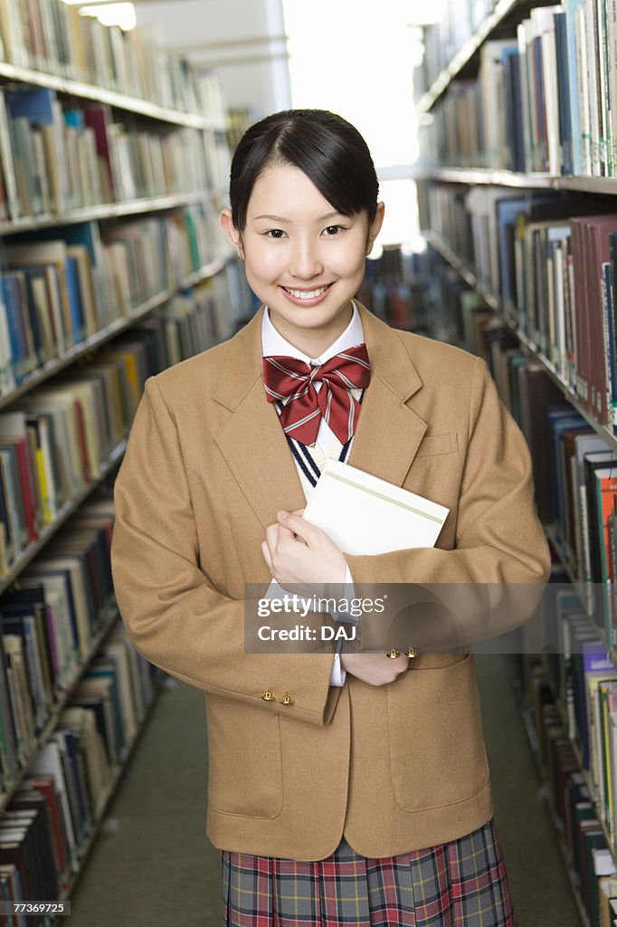 Portrait of a teenage girl holding a book in library, smiling and looking at camera
