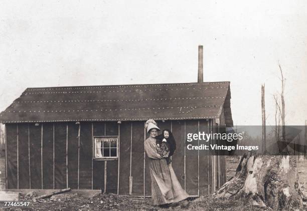 Woman holds an infant in her arms as she poses in front of a claim shack, early 1910s. The structure, mostly walled in tar paper, served to lay claim...