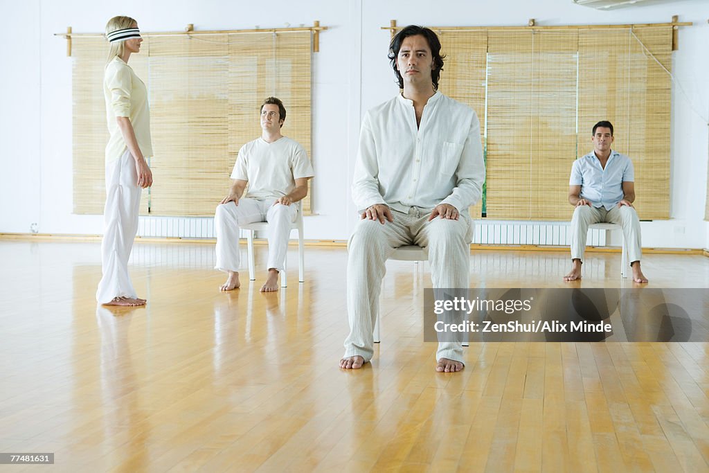 Group therapy, adults sitting in chairs while one woman stands wearing blindfold