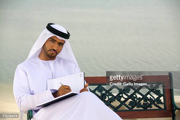 arab man sitting on park bench writing journal. dubai, united arab emirates - arabische muster stockfoto's en -beelden