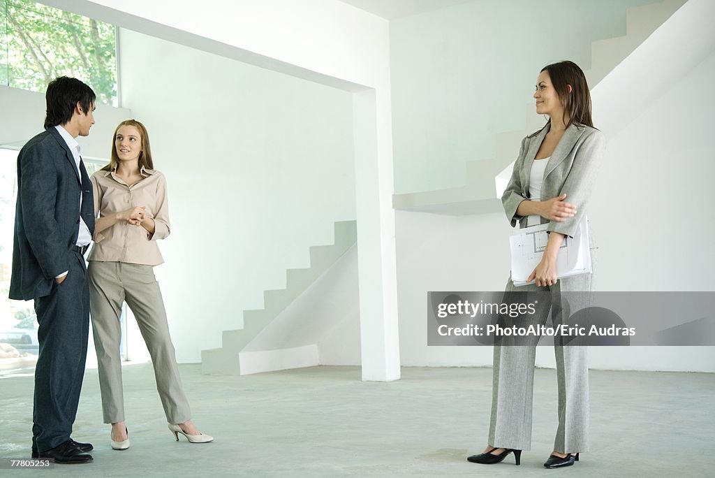 Female real estate agent standing in empty home interior with young couple, full length