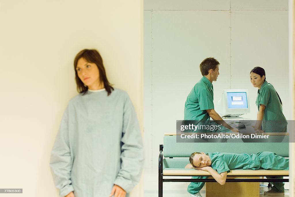 Medical workers chatting in office, one lying down on bench, female patient standing in foreground