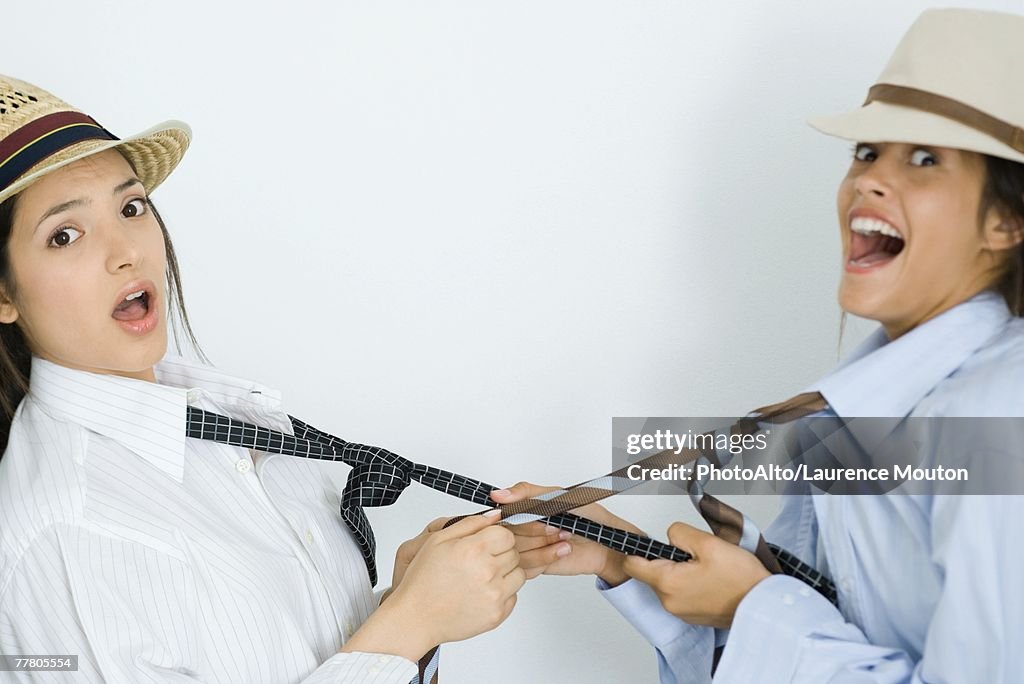 Two young female friends dressed in hats, pulling each other's ties, looking at camera