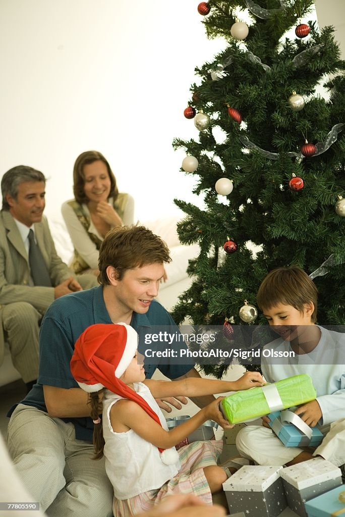 Father and two children sitting by Christmas tree, opening presents together, adults in background