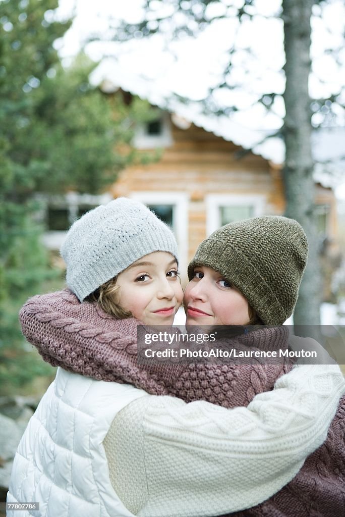 Two young friends embracing, dressed in winter clothing, looking over shoulders at camera, portrait