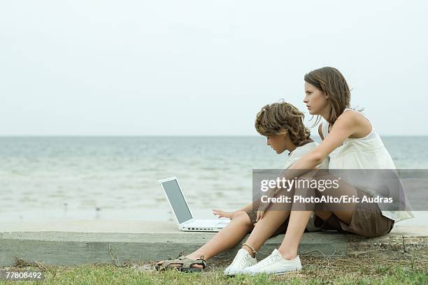 teen girl and little brother using laptop computer together by ocean, side view - teen boy shorts stockfoto's en -beelden