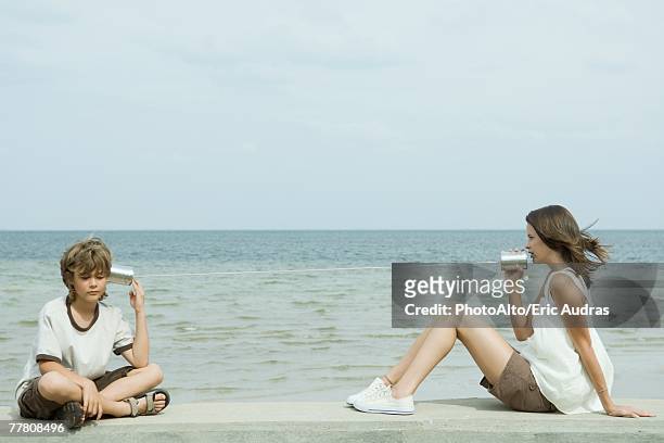 boy and teen sister sitting next to water, talking to each other through tin can phone - teen boy shorts stockfoto's en -beelden