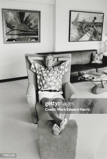 Portrait of German-born Australian photographer Helmut Newton as he sits in a amrchair, his hands behind his head and his bare feet up on a stool, in...