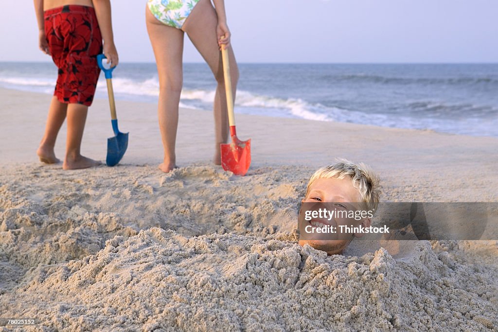 A boy smiles to the viewer while buried in the sand, and two kids stand with shovels in the background