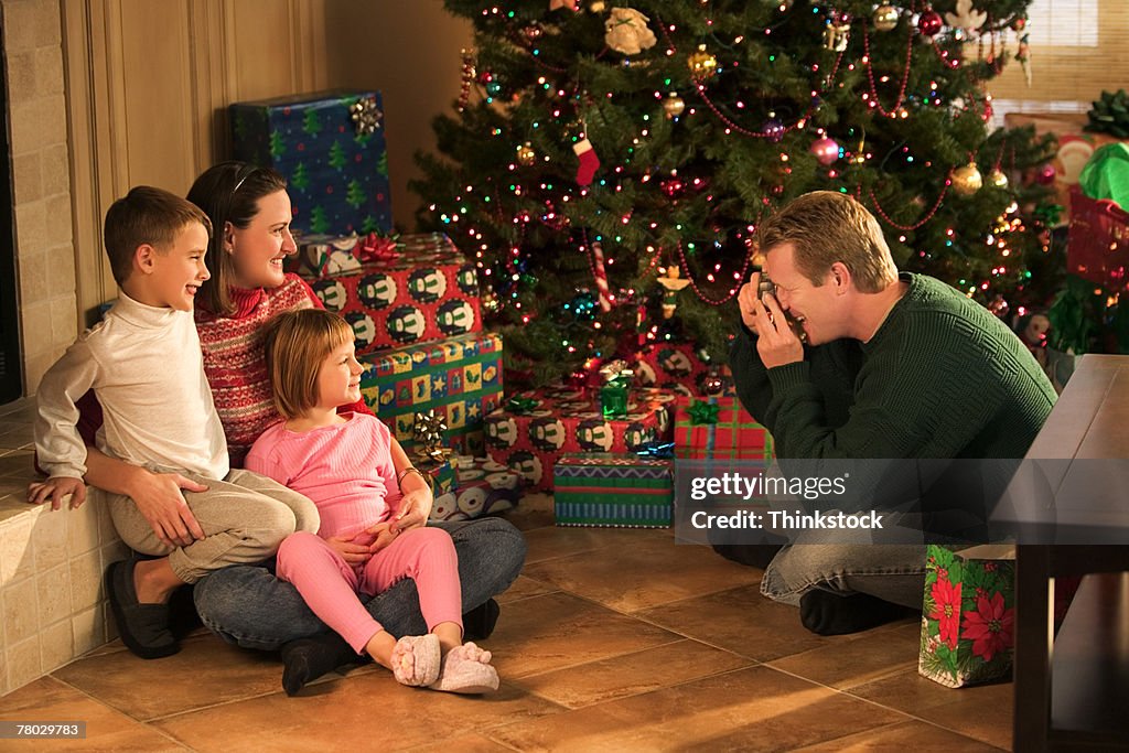 A father takes a photo of his wife and children near the Christmas tree.