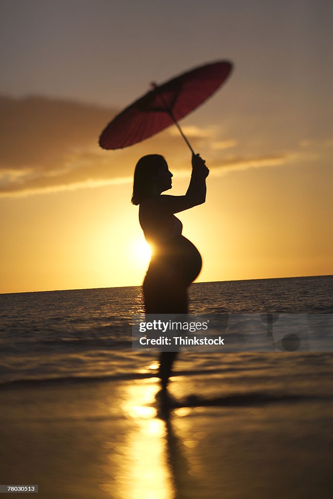 Backlit profile of a pregnant woman standing on the beach at sunset holding a red parasol above her head.