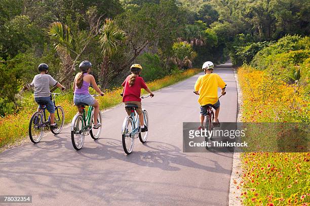 rear view of four kids riding bicycles on a small road away from the viewer - teenager cycling helmet stock pictures, royalty-free photos & images