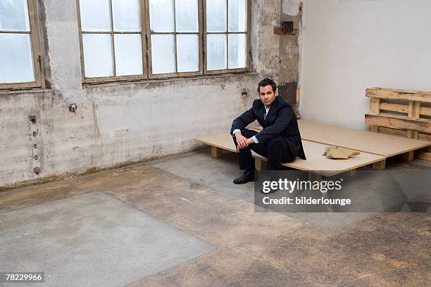 businessman sitting on wooden pallet in empty building - unhappy salesman stock pictures, royalty-free photos & images