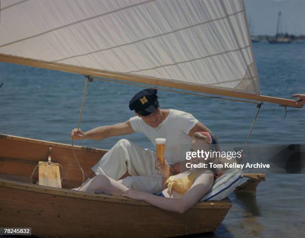 Young woman in a yellow bikini top holds up a tall glass of beer as she leans back in a small sailboat, watched intently by a man in a captain's cap...