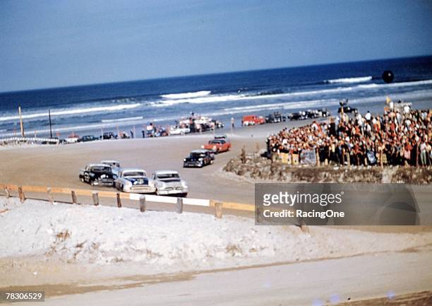 Blue Atlantic Ocean waves break on the shoreline as Grand National stock cars slam their way around the North Turn on Daytona's beach race course in...