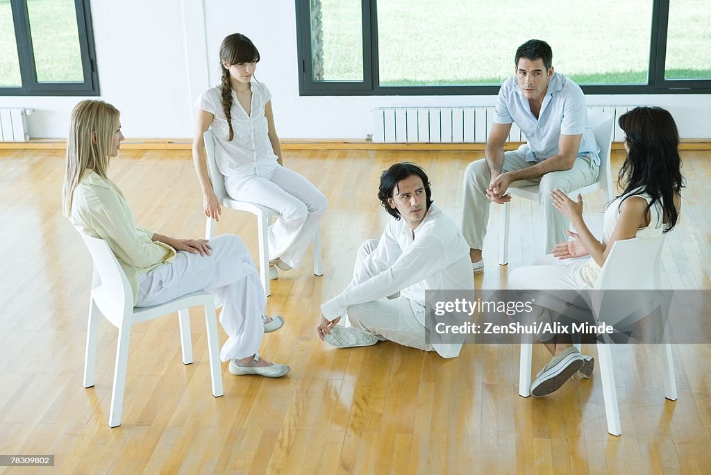 Man sitting on floor in middle of group sitting in chairs, talking