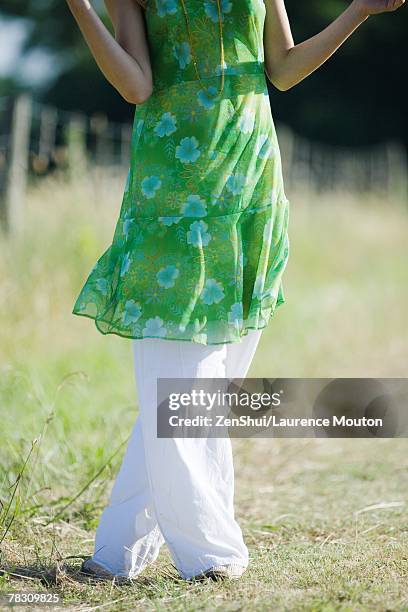 young woman standing in rural field with legs crossed at knees, cropped view - tuniek stockfoto's en -beelden