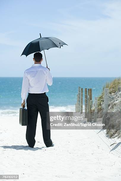 businessman standing under umbrella on sandy path leading to ocean, rear view - out of context stock pictures, royalty-free photos & images