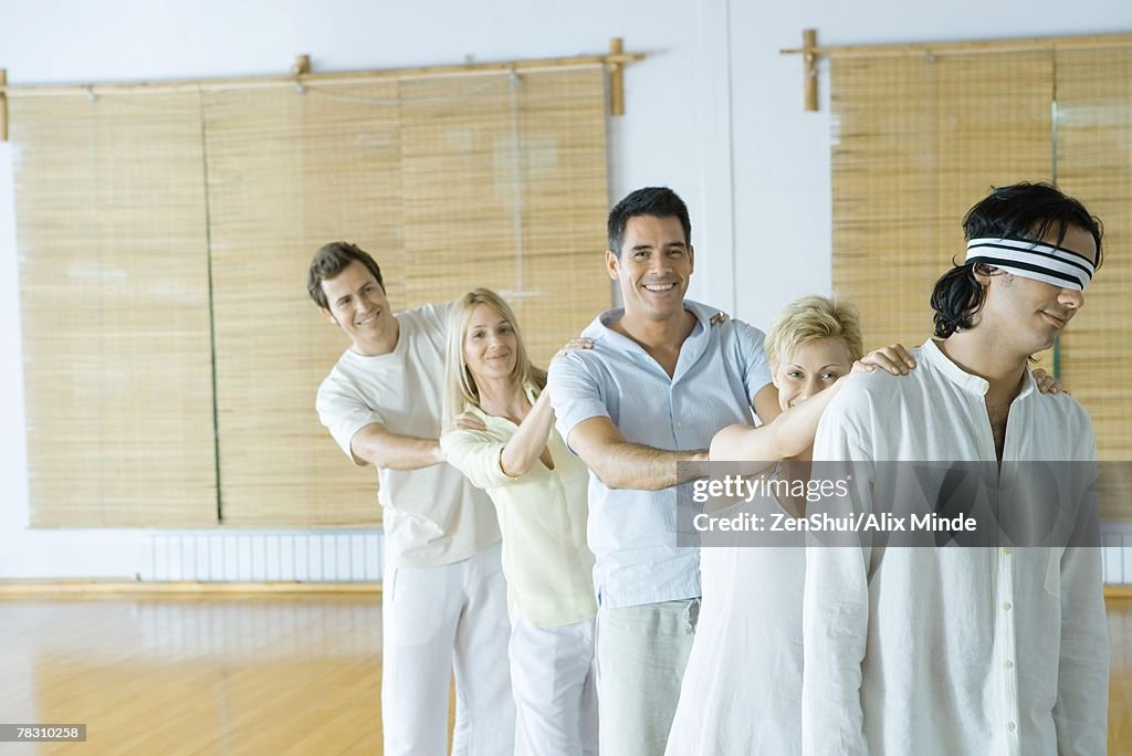 Group of adults standing single file holding onto each others' shoulders, leader blindfolded